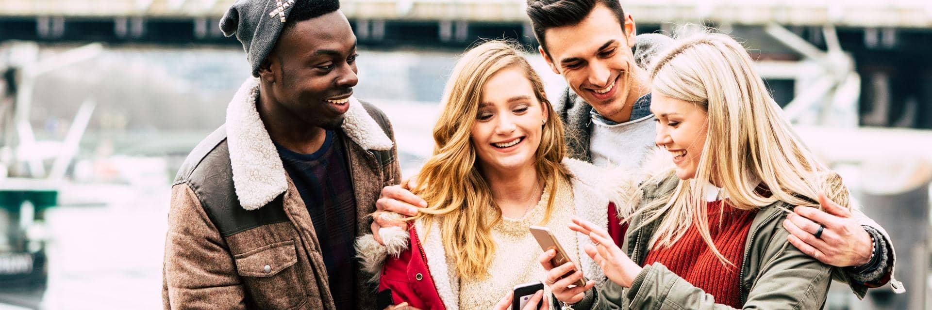 A group of young people wearing winter clothes laugh as they look at something on a mobile phone