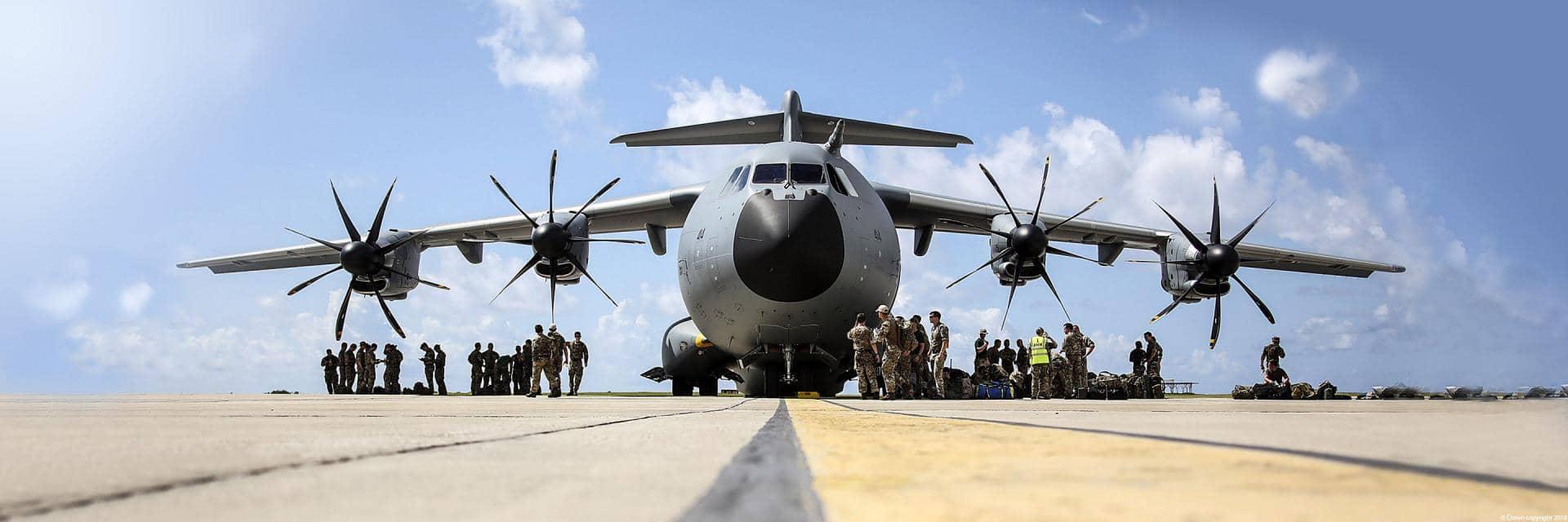 A military airplane on a runway with troops and kit assembled under both wings
